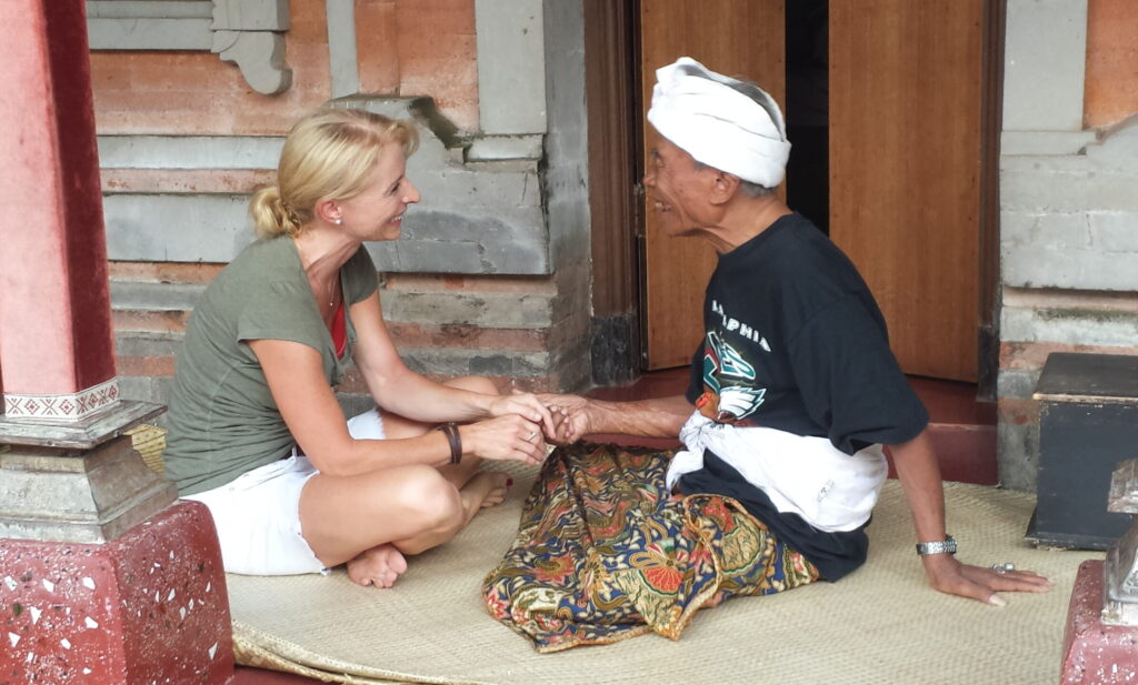 Christina Soltau im fröhlichen Austausch mit Ketut Lear - einem alten, sehr bekannten Heiler aus Bali. Beide sitzen im Schneidersitz auf dem Boden. Ketut hält Christinas Hand.