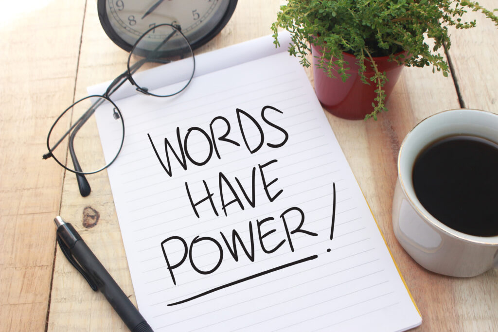 "Words have power!" auf einem Block geschrieben. Daneben eine Tasse Kaffe, eine Brille, Stift und Pflanze